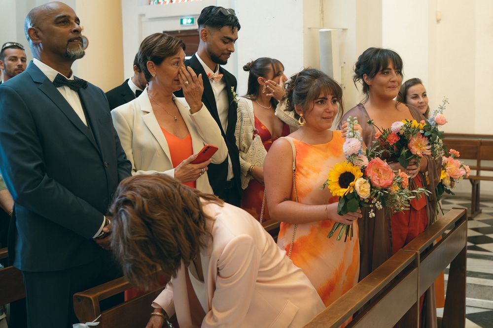 Wedding guests in orange dresses watch ceremony from church pews.