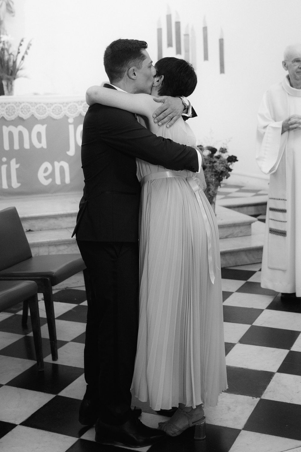 Black and white photo of a couple embracing on a checkered floor at a celebration.