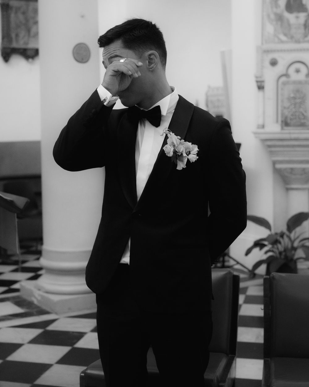 A person wearing a formal black suit adjusts their tie in an elegant setting.