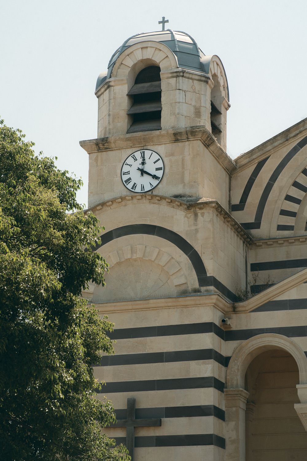 Church tower with striped architecture and clock face against pale blue sky.