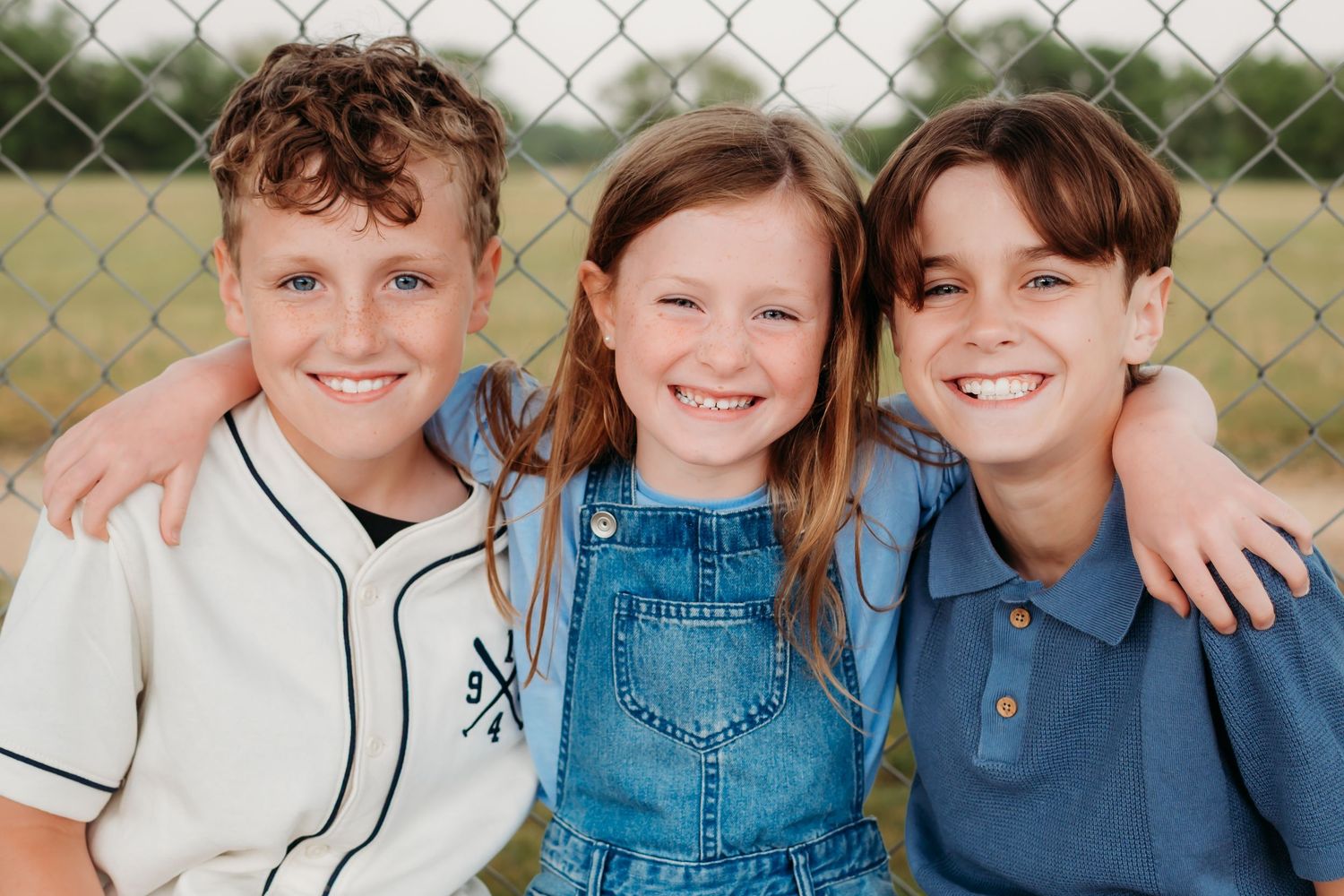Children in baseball uniforms and casual clothing sit side by side on a fence outdoors in a cheerful group photo.