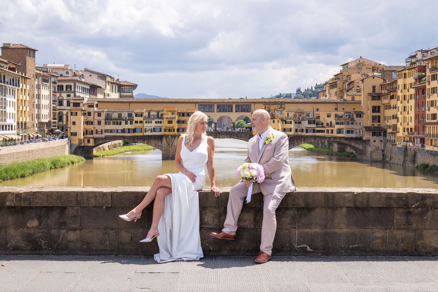 Couple in elegant white outfits sitting on stone wall overlooking the historic Ponte Vecchio bridge in Florence, Italy.