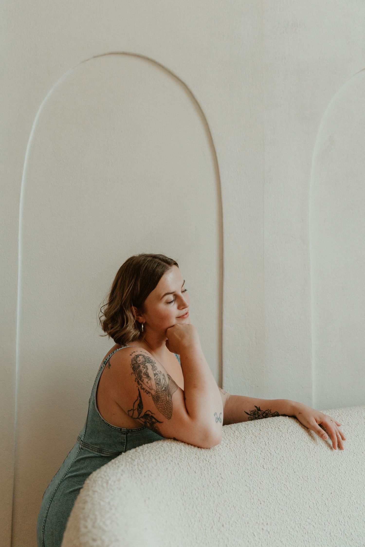 A person in a sage green dress sits thoughtfully by a white arched wall, showing an artistic arm tattoo.