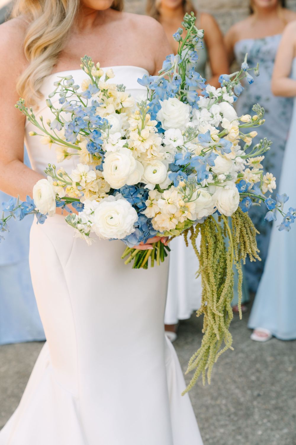 Bride holding a lush bouquet of white ranunculus and blue delphinium flowers with bridesmaids behind her.