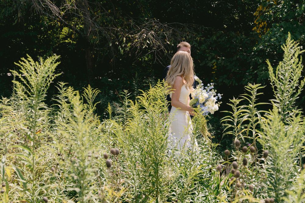 Bride in white lace gown holding bouquet stands among tall wildflowers in a sunlit garden.