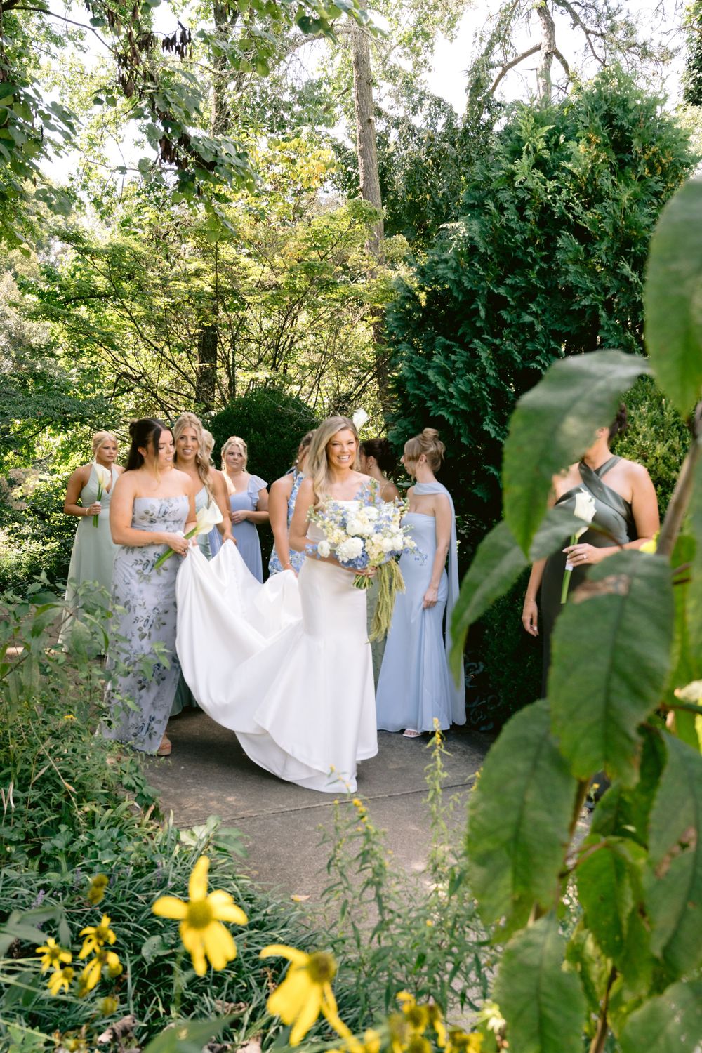 Bride in white gown holds bouquet surrounded by bridesmaids in blue dresses in lush garden setting.