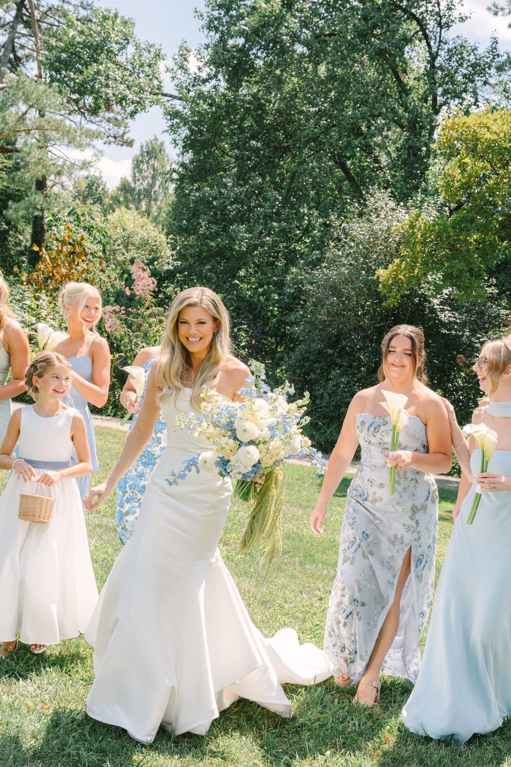 Bride in white gown walks with bridesmaids in blue dresses and flower girl on sunny lawn.