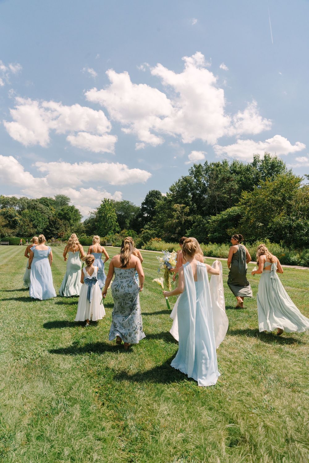 Bridesmaids in light blue dresses walk across a sunny green lawn with a bride in white at an outdoor wedding.