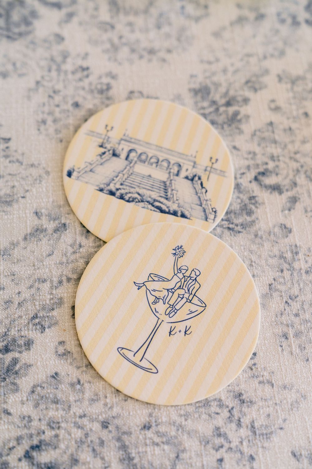 Two round yellow striped wedding coasters featuring blue ink illustrations of a couple in a martini glass and a venue.
