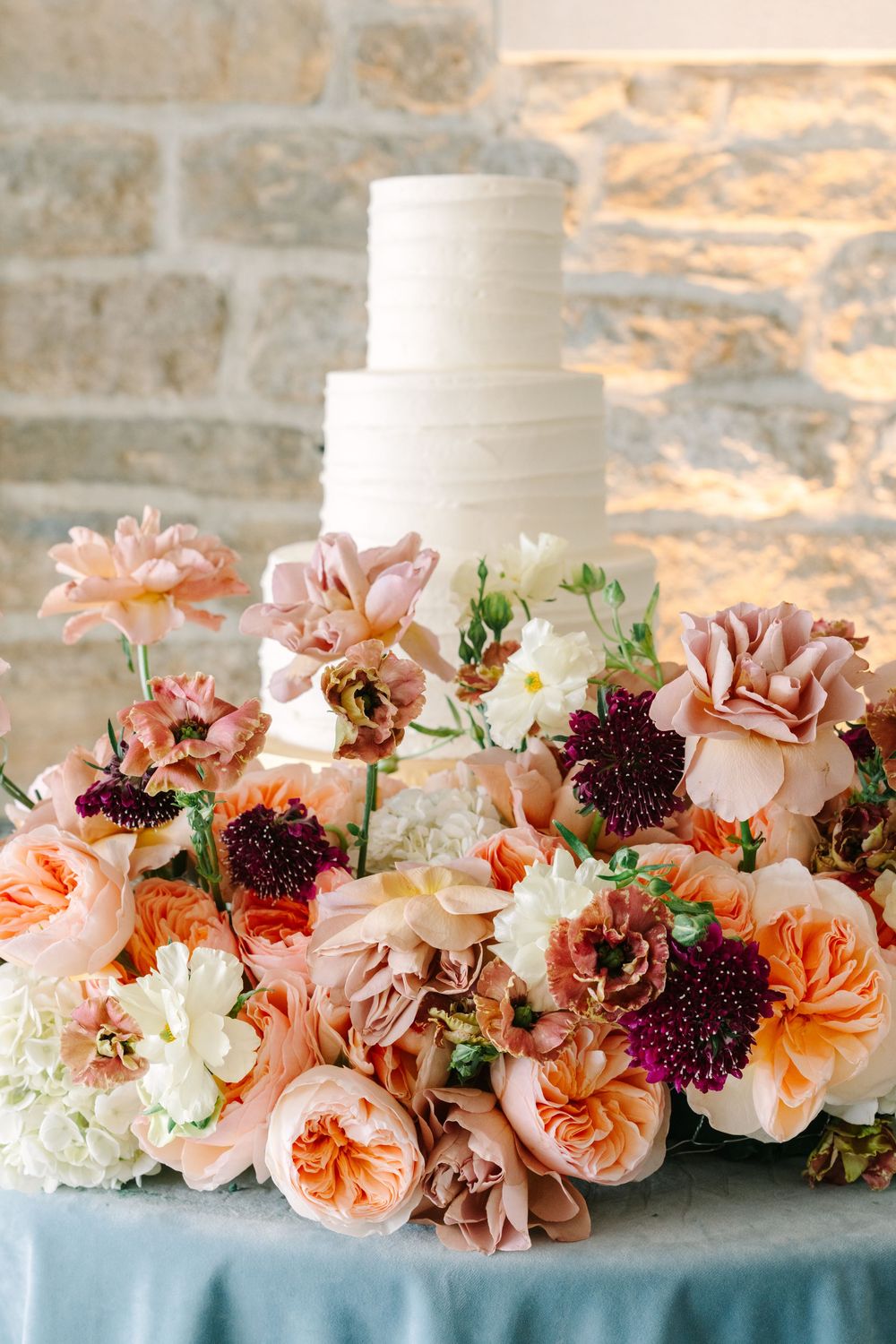 Elegant three-tier white wedding cake surrounded by peach, pink, and burgundy flowers on a blue tablecloth.