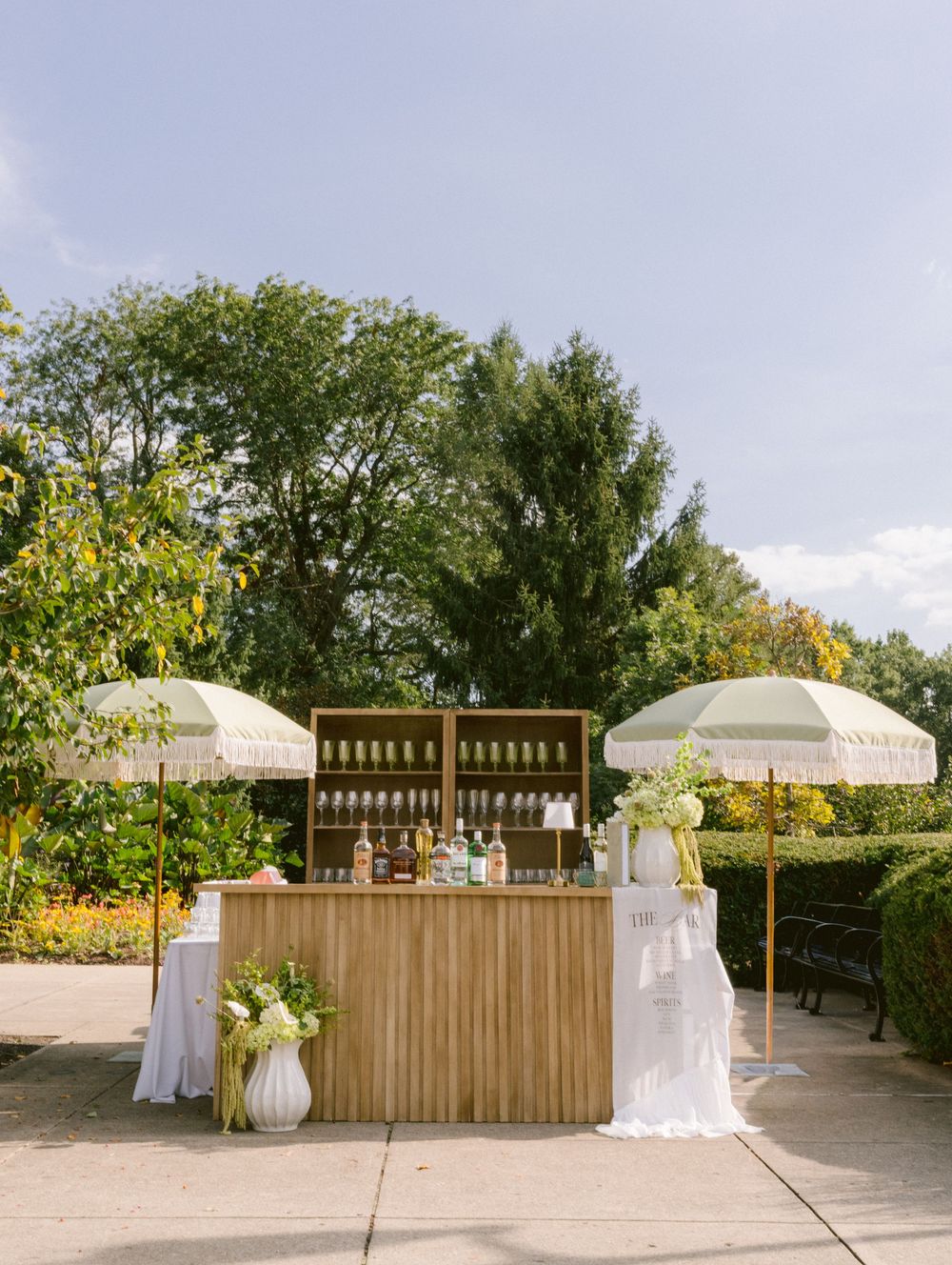 Elegant outdoor wedding bar with wooden counter, striped umbrellas, and floral arrangements on a sunny day.