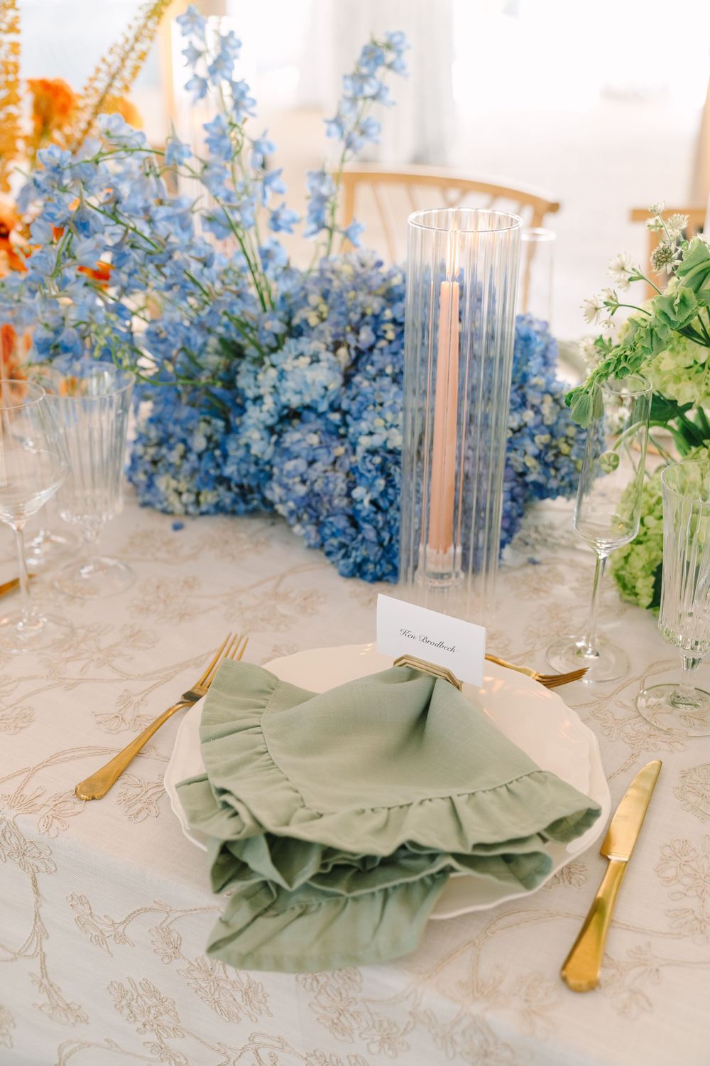 Blue hydrangea wedding tablescape with sage green ruffled napkins, gold cutlery, and tall taper candles.