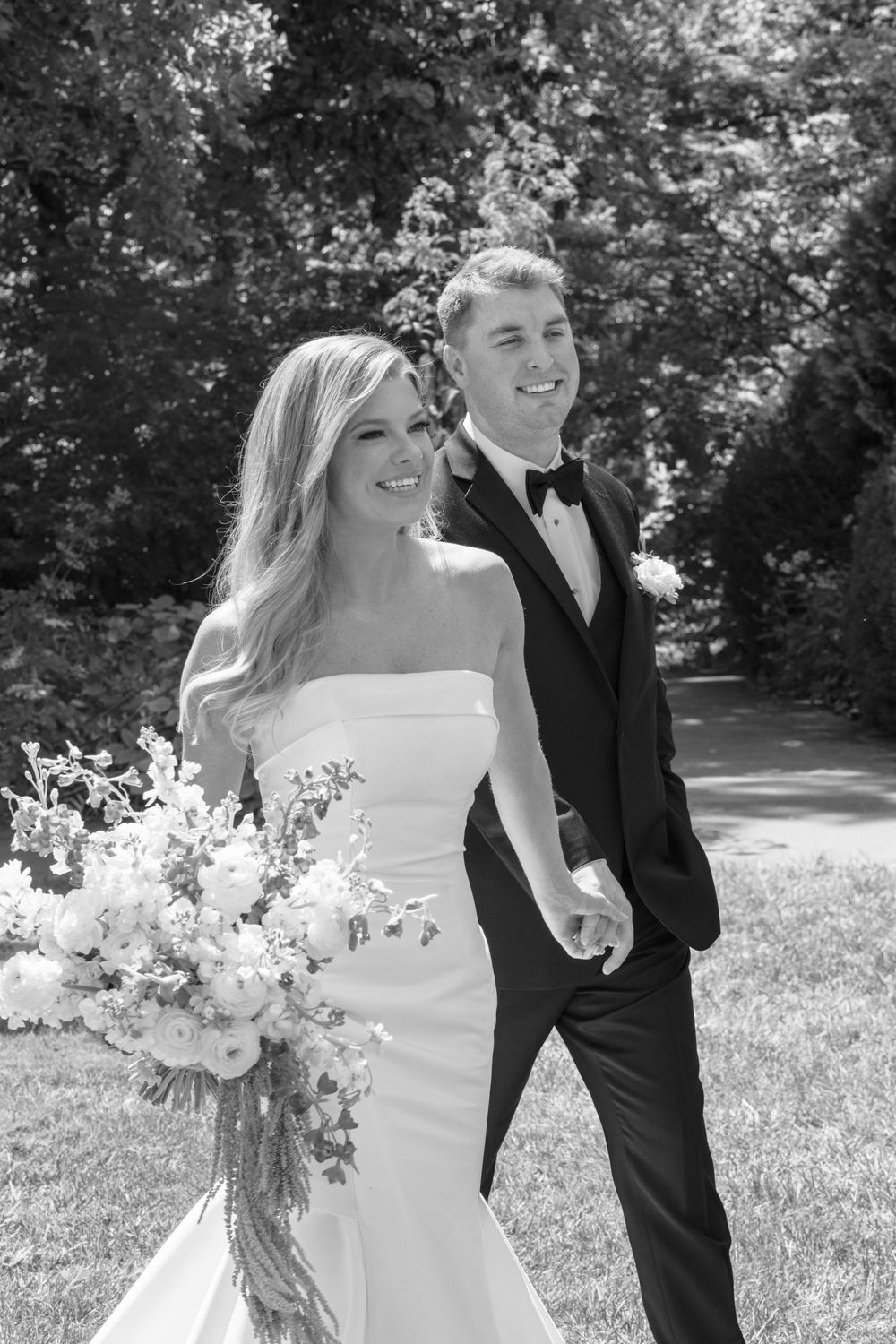Black and white photo of smiling bride holding bouquet and groom in tuxedo walking outdoors on their wedding day.