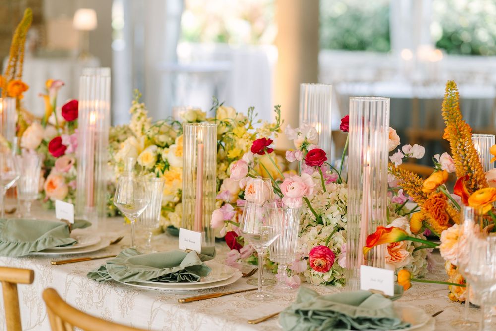 Elegant wedding reception table with colorful floral arrangements in pink, yellow, and orange, tall glass candles, and sage green napkins.