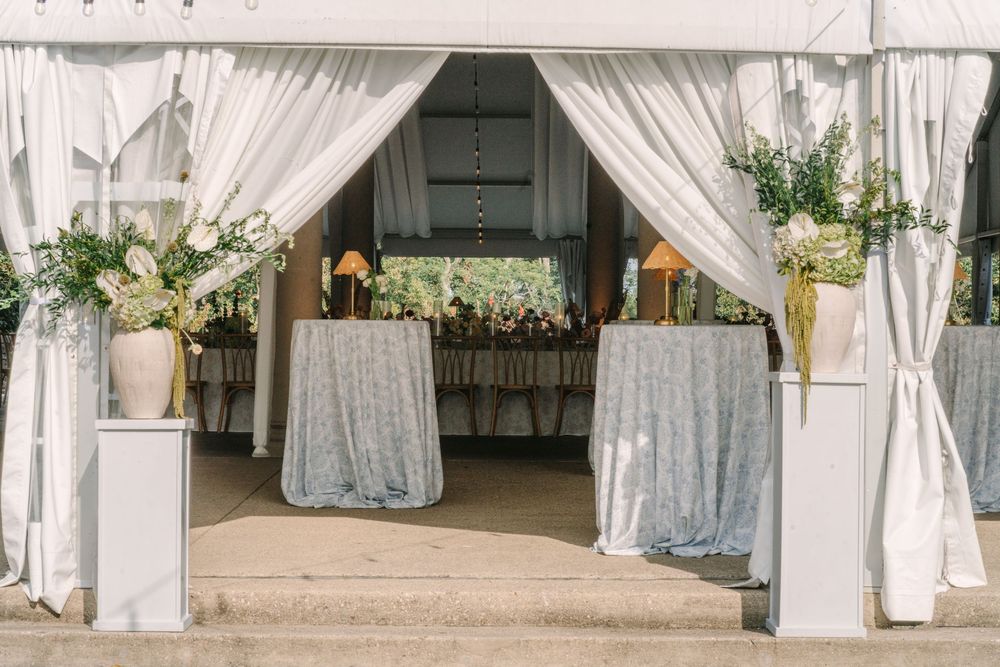 Elegant wedding tent entrance with white draping, floral arrangements, and silver tablecloths creating a grand reception entry.
