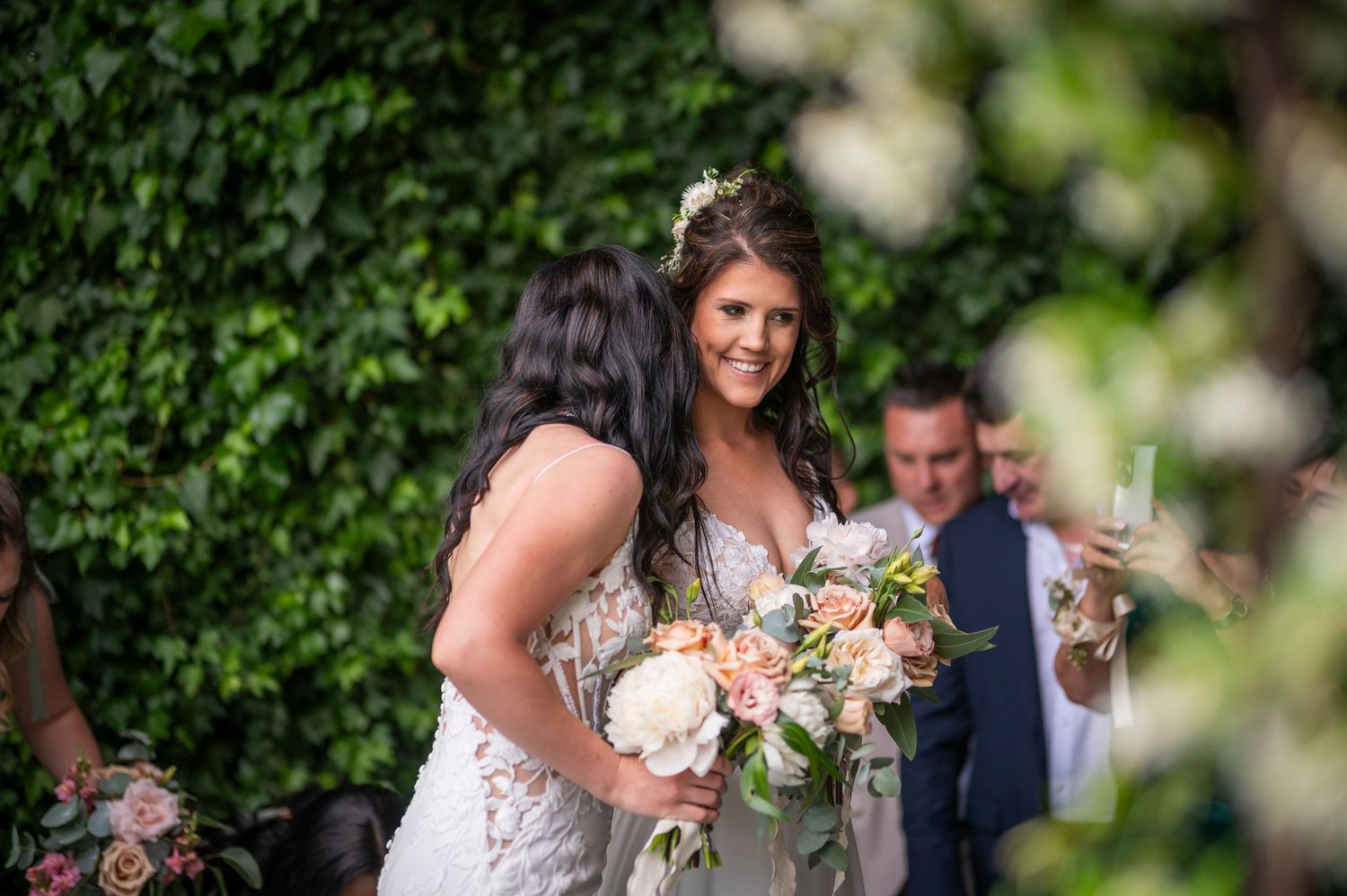 A bride in a fitted white lace gown holds a romantic pastel bouquet while standing in front of a lush ivy wall.
