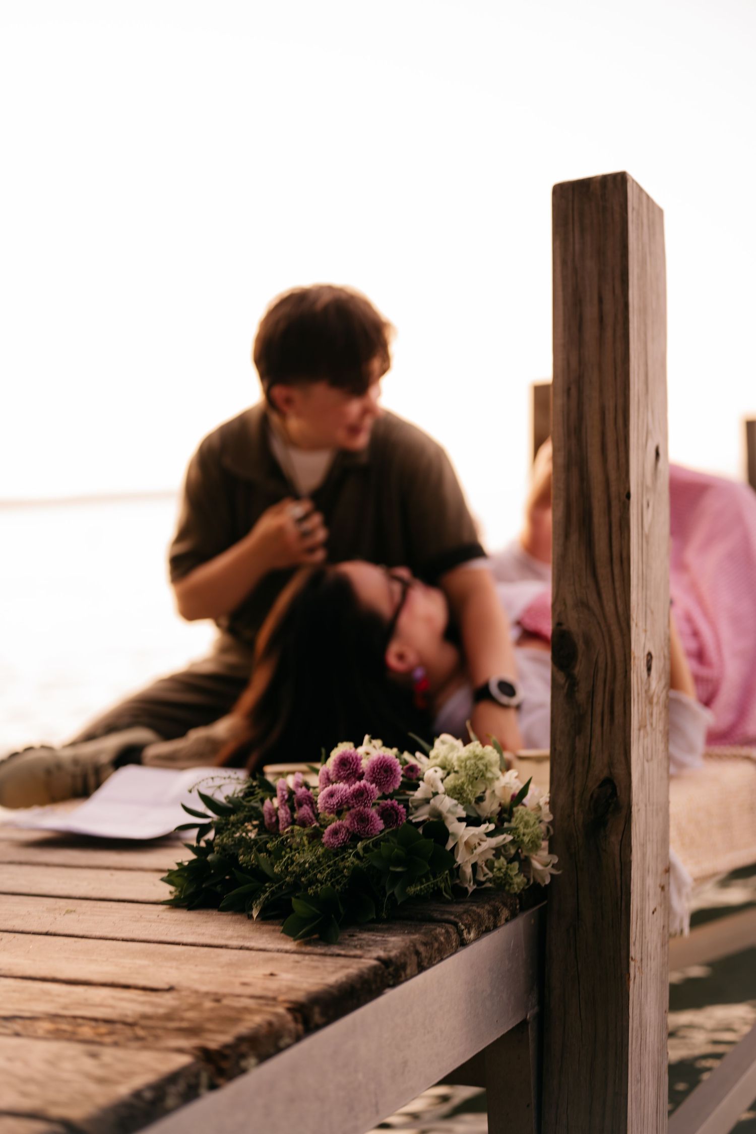 A couple relaxes on a wooden dock at sunset with a floral bouquet in the foreground.