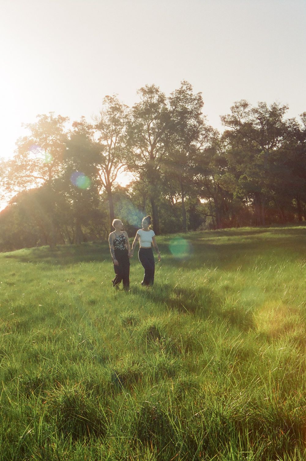 Queer couple anniversary photoshoot in Perth, Sarah and Viviane walking through a sunlit field, LGBTQ+ friendly photography.