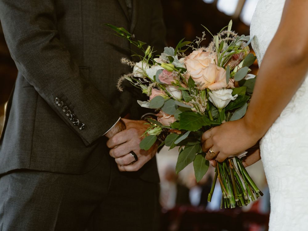 A peach and white floral wedding bouquet being held between two formal attire-clad hands.