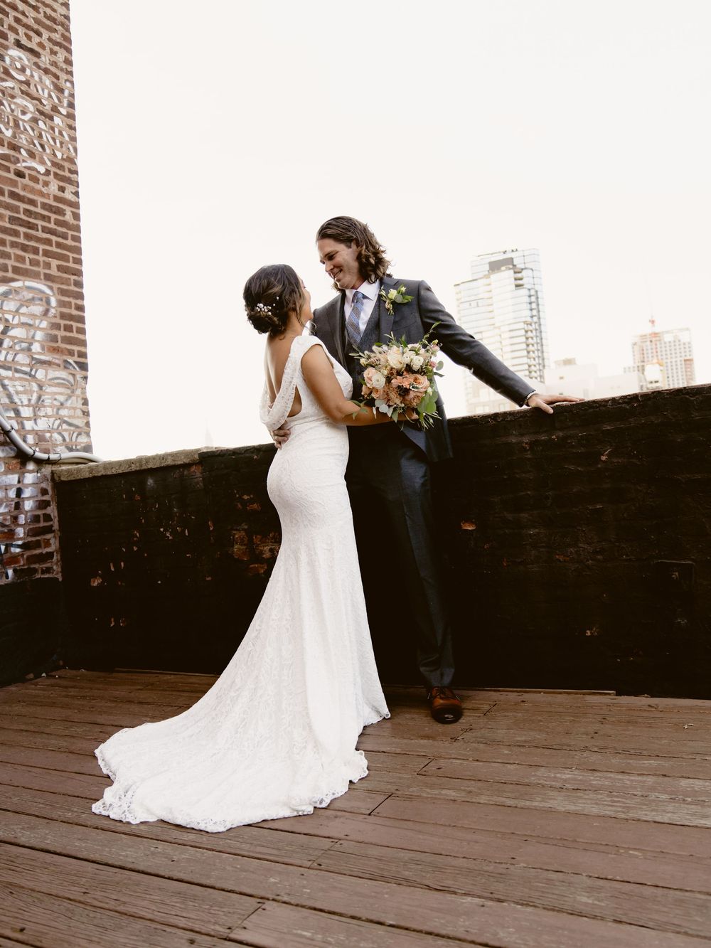 An elegant wedding couple on a rustic rooftop with a trailing white dress and urban backdrop.