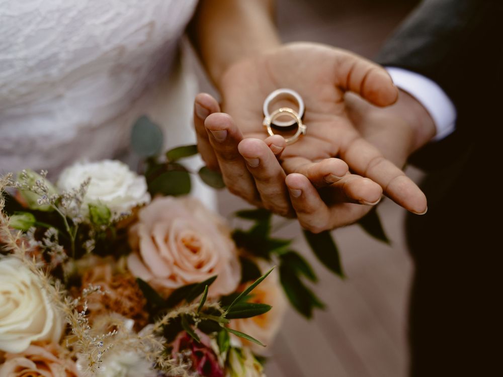 Wedding rings displayed in open palms with a soft focus floral bouquet in the background.