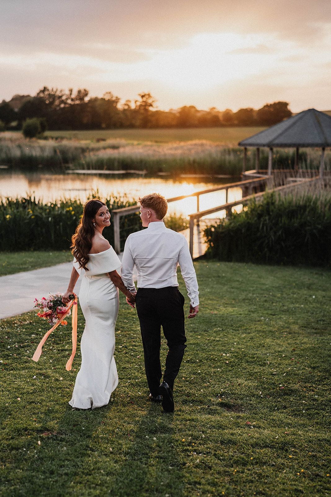 A couple in wedding attire walks together on grassy fields during golden hour sunset beside a lake.