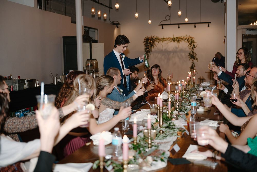 Wedding reception guests enjoy dinner at long tables decorated with greenery and candlelight under pendant lighting.