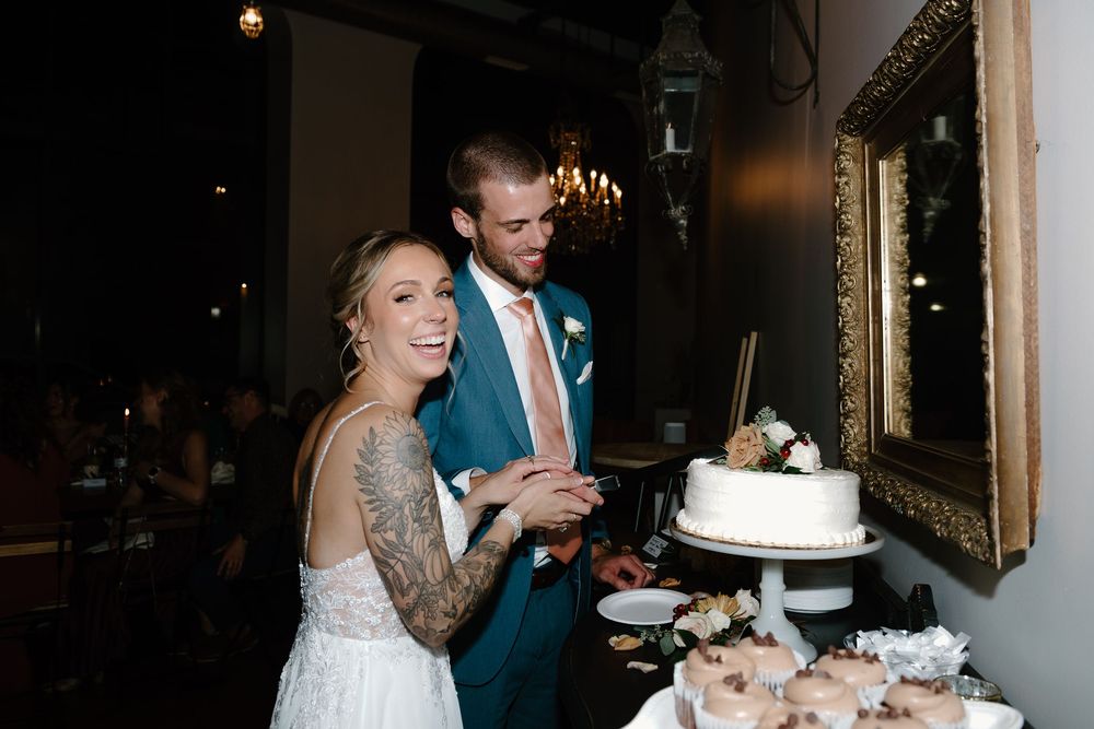 Couple shares a joyful moment while cutting their elegant white wedding cake together at an indoor reception.