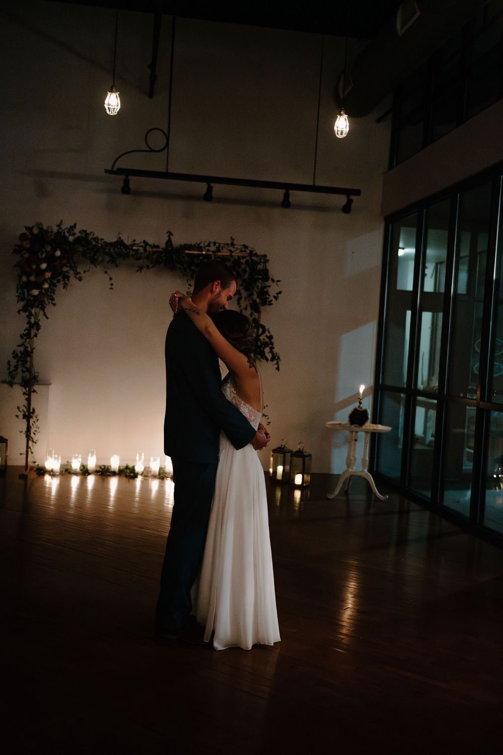 A couple shares their first dance in a dimly lit venue decorated with candles and floral archway.
