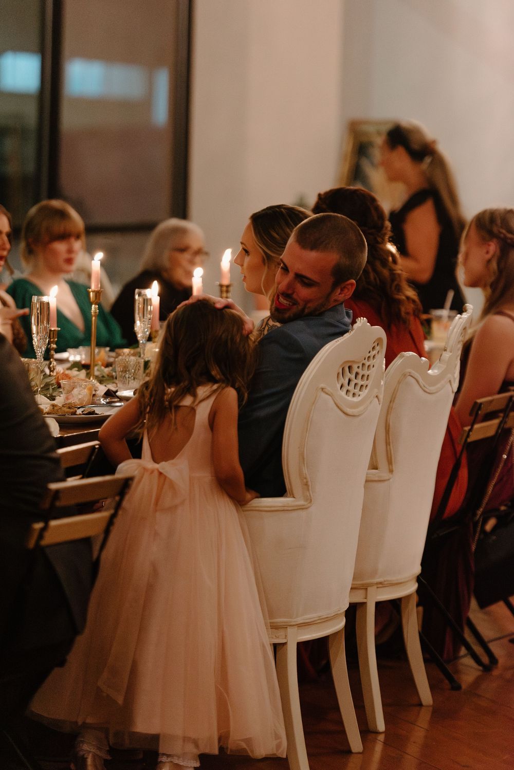 Guests seated at an elegant wedding reception dinner with white chairs and candlelit tables in a dimly lit venue.