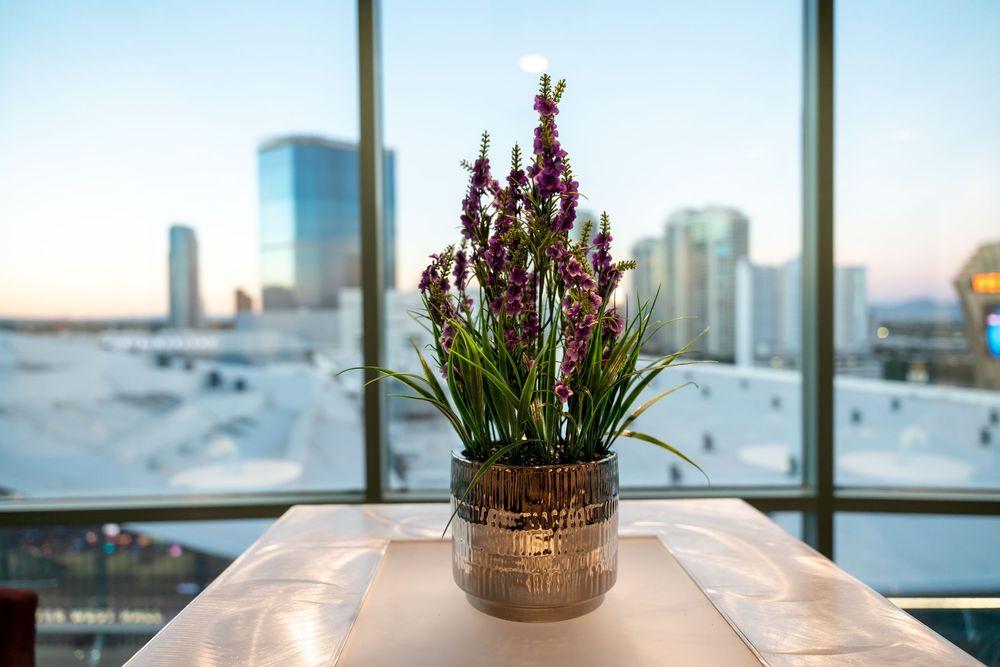 Purple flowers in a decorative vase sit by a window overlooking a modern city skyline at sunset.