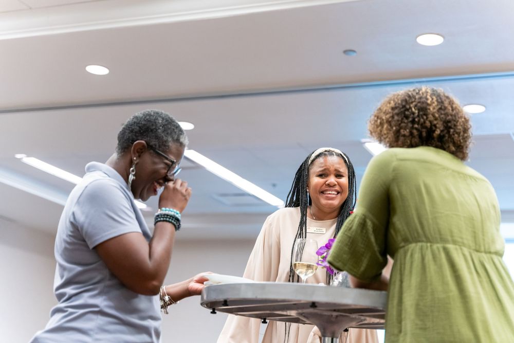 Three people engage in animated conversation around a table in an office space with recessed lighting.