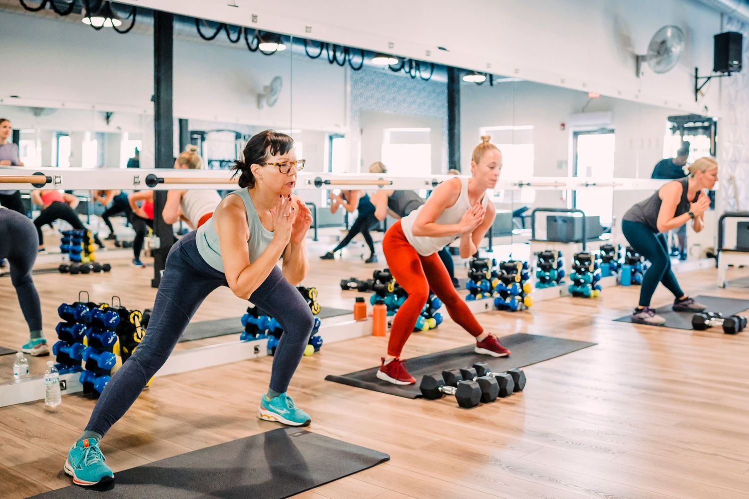 Group fitness class participants performing lunges with step platforms and weights in a bright studio gym.