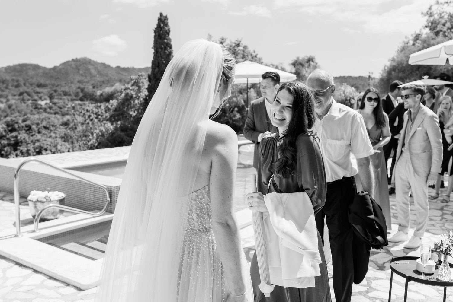 A black and white wedding photograph showing a bride in a veil greeting guests at an outdoor reception with mountains in view.