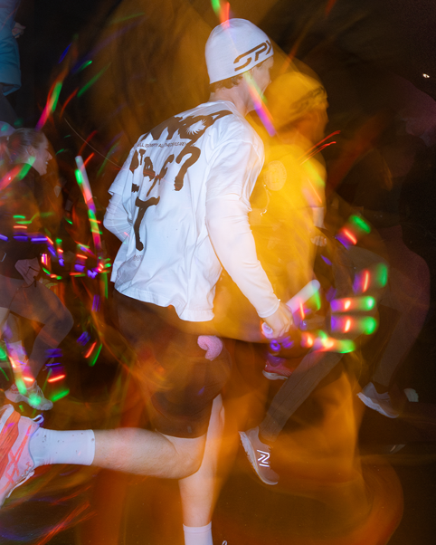 Energetic runner in white shirt captured in motion with colourful light trails and effects at a night time event.