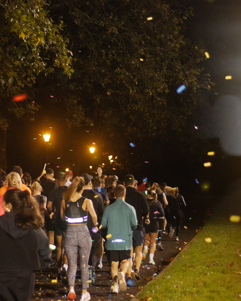 A nighttime running group follows path lit by street lamps, with colourful lights illuminating the trees overhead and confetti raining down.