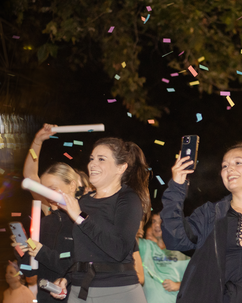 Enthusiastic participants celebrating with light sticks and recording moments on their phones as confetti falls during night time event.