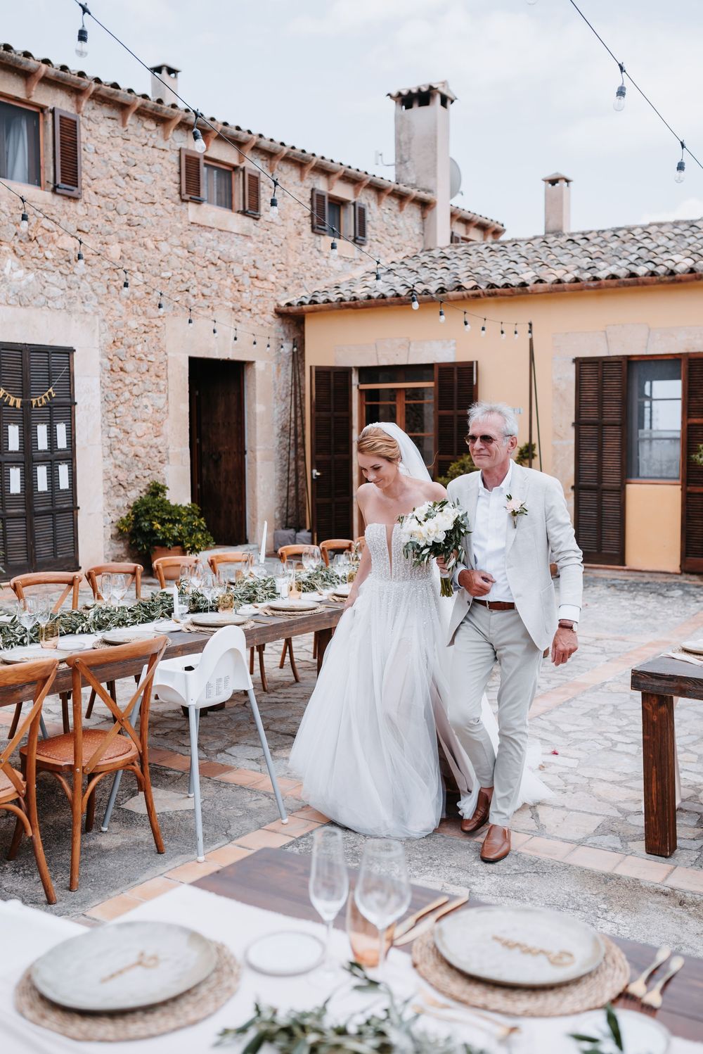 Der Brautvater führt seine Tochter zur Zeremonie zwischen eleganten, mediterranen Holztischen des Hochzeitsdinners und Steinarchitektur.