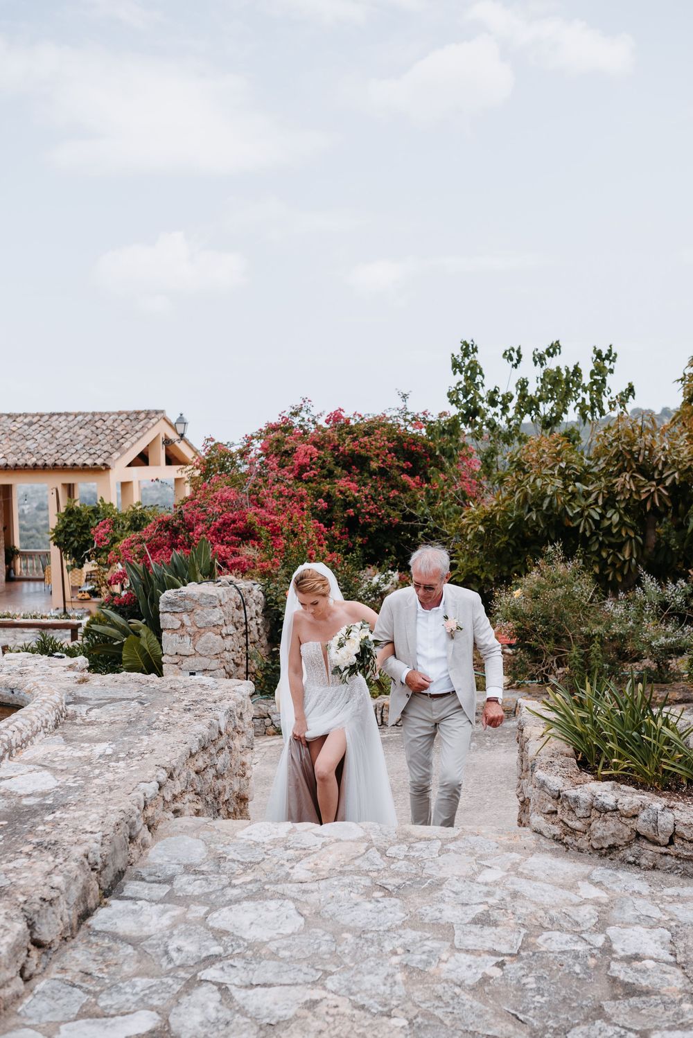 Einzug des Brautvaters mit seiner Tochter. Sie gehen auf einem Steinweg, umgeben von rosa Bougainvillea und mediterraner Finca Architektur.