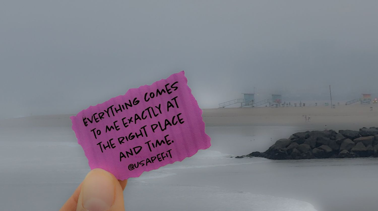 A pink sticky note with motivational text is held up against a foggy beach landscape in the background.