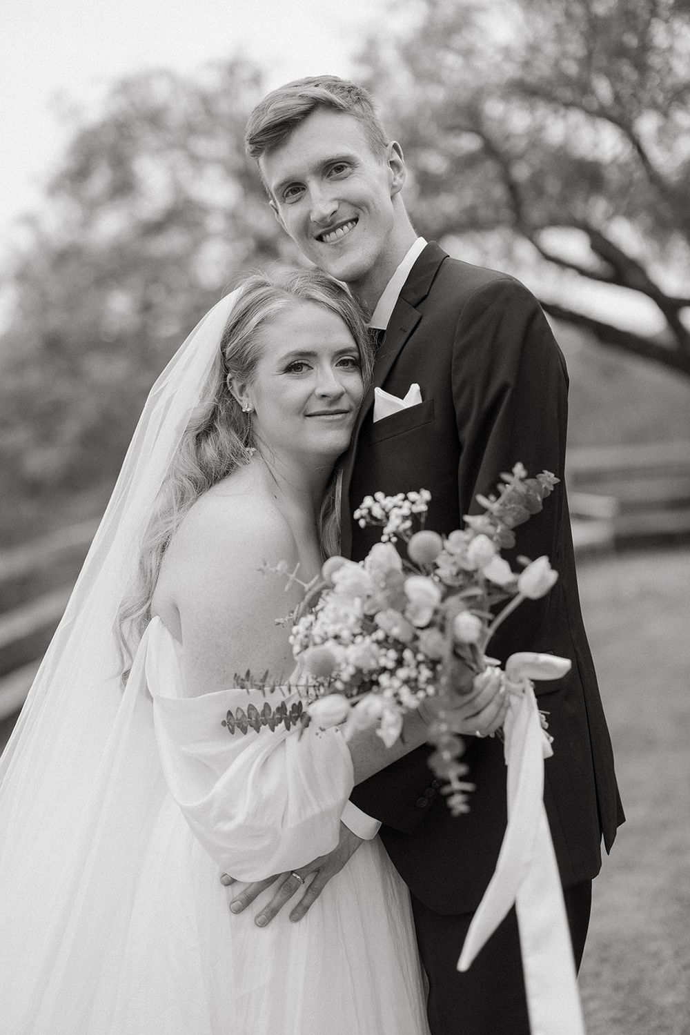 Black and white photo of newlywed couple outdoors, bride wearing veil and holding floral bouquet, groom in formal suit.