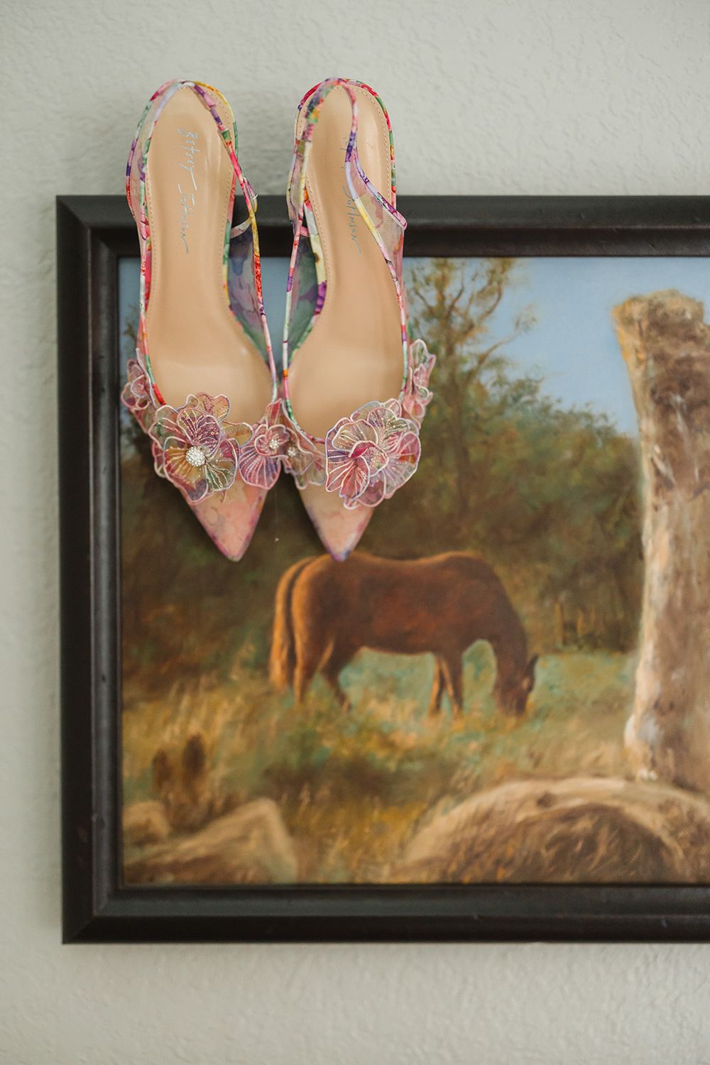 A pair of pink floral embellished shoes displayed against a wall with a landscape painting of horses in the background.