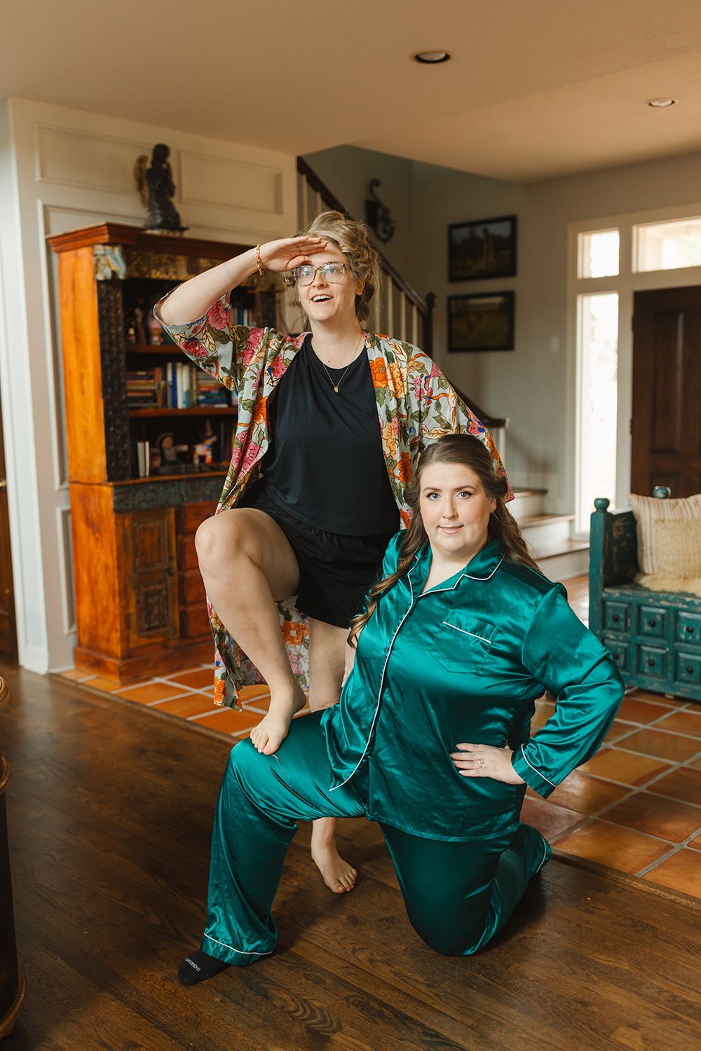 Two people pose playfully in casual loungewear, one in a floral robe and one in emerald satin pajamas, on hardwood floors at home.