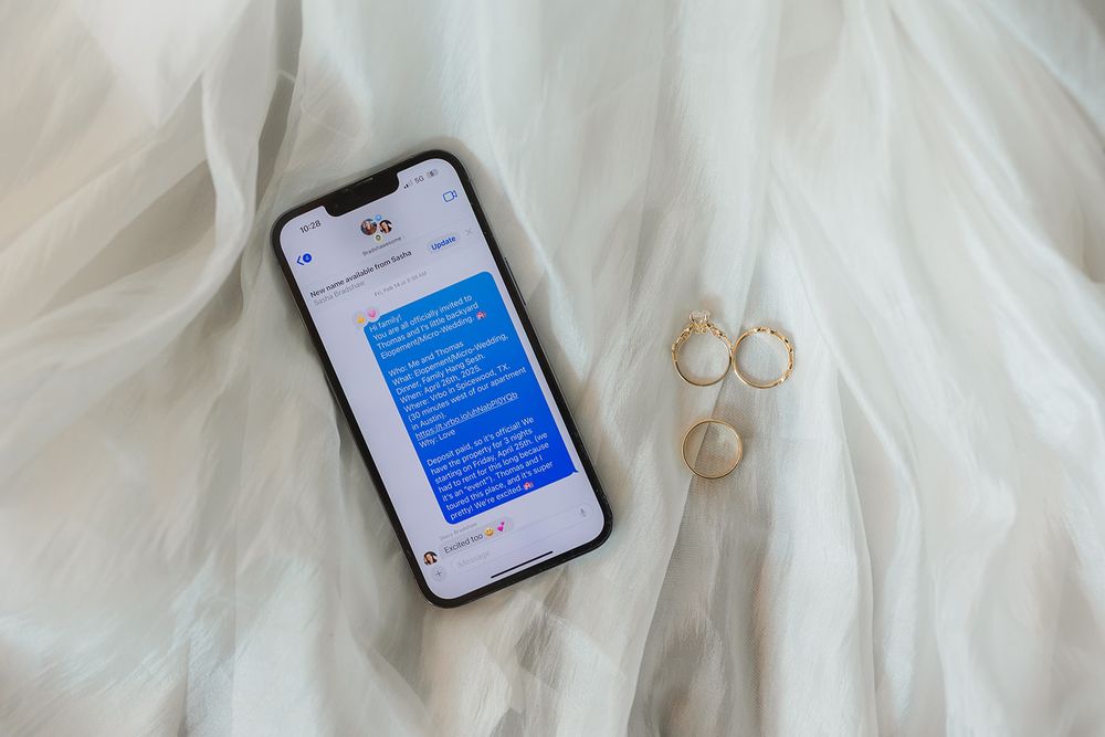iPhone displaying blue text messages lies on white fabric next to gold rings and jewelry.