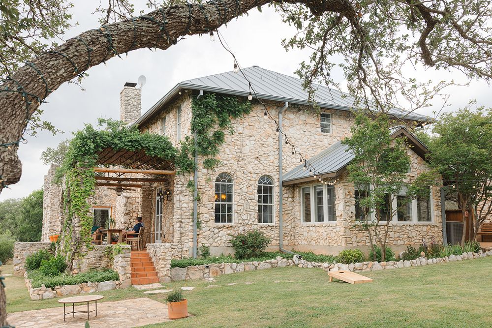 Rustic stone house with ivy-covered walls and arched windows sits beneath a sprawling tree branch on a manicured lawn.
