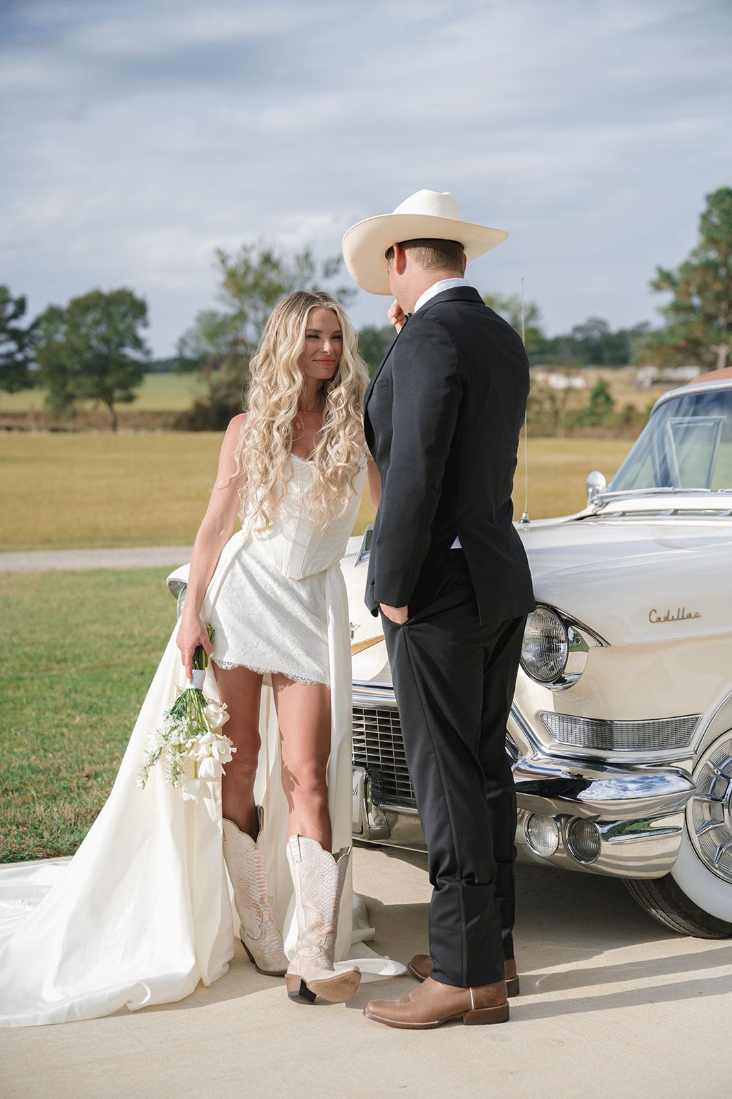 Couple stands by vintage Cadillac at country wedding with bride in short dress with train and groom in cowboy hat.