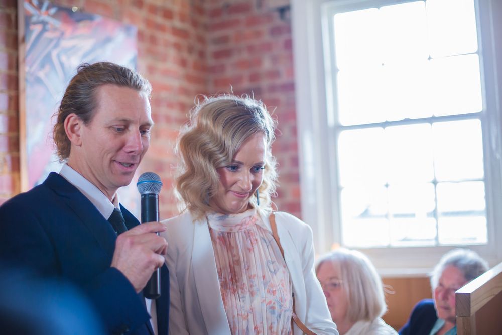 Two people speak into a microphone at an indoor event with exposed brick walls and natural lighting through windows.