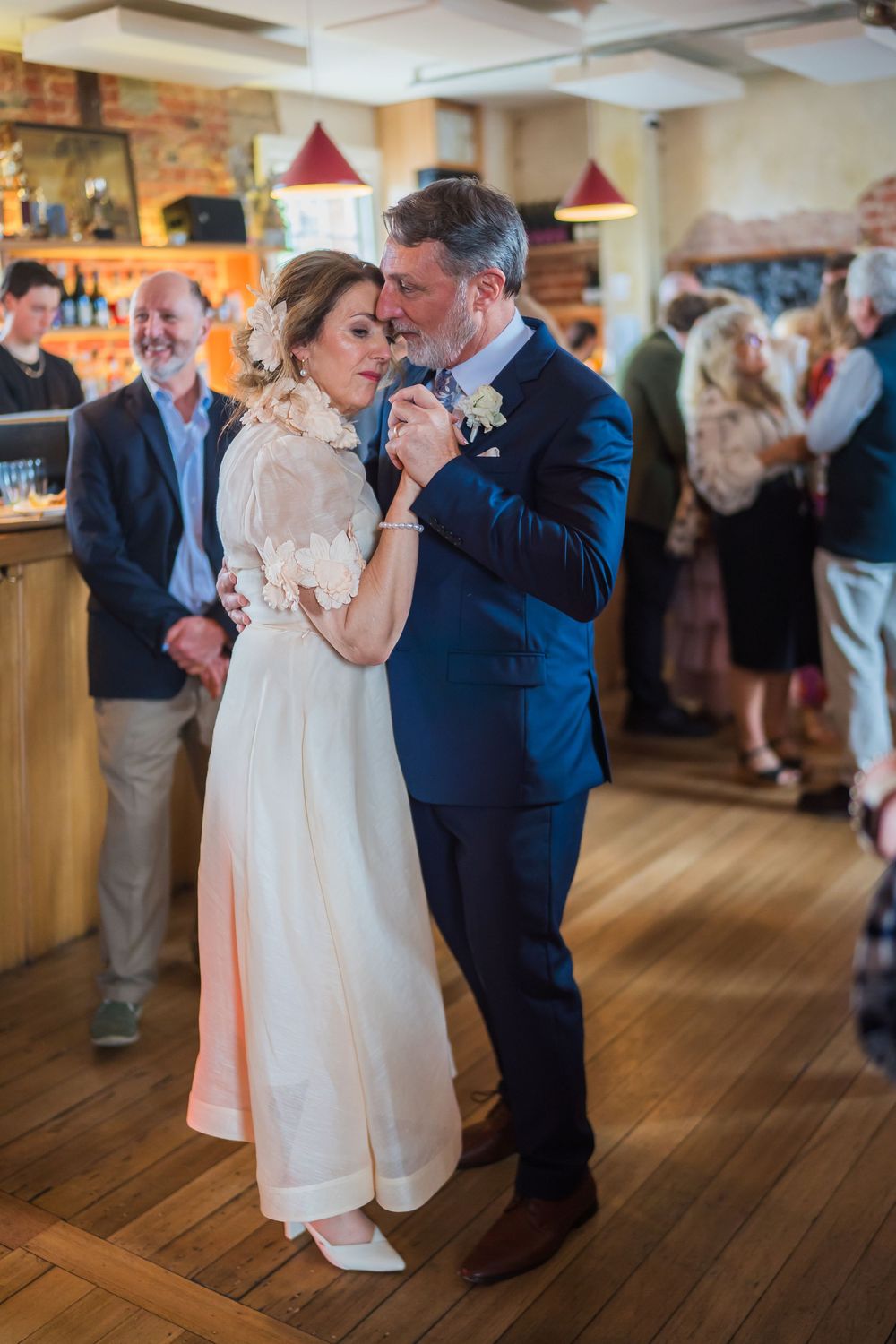 Newlyweds share an intimate first dance on wooden floor at wedding reception with guests looking on in background.
