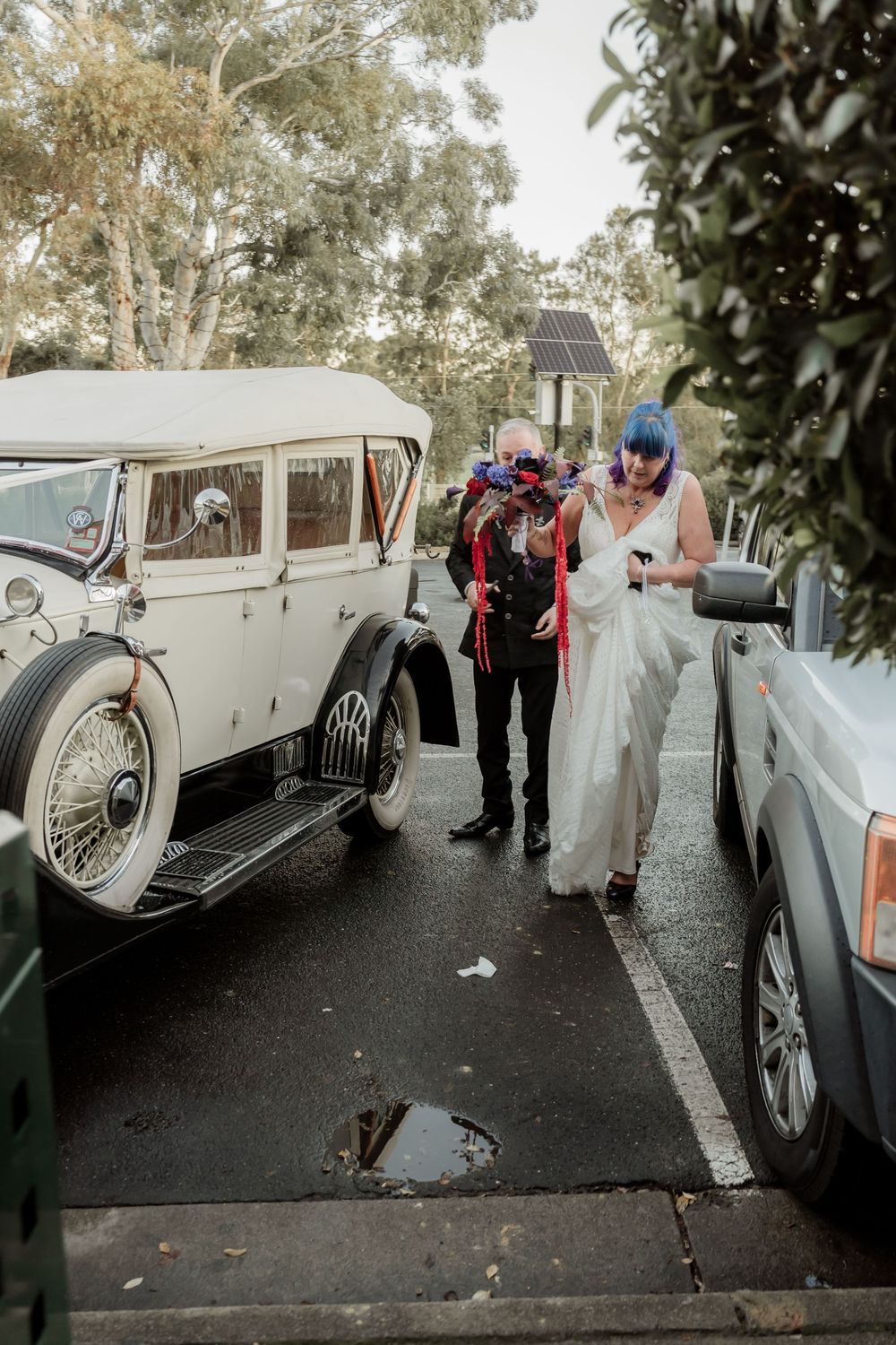 Vintage white classic car parked between modern vehicles with a couple dressed in formal attire at an outdoor event.