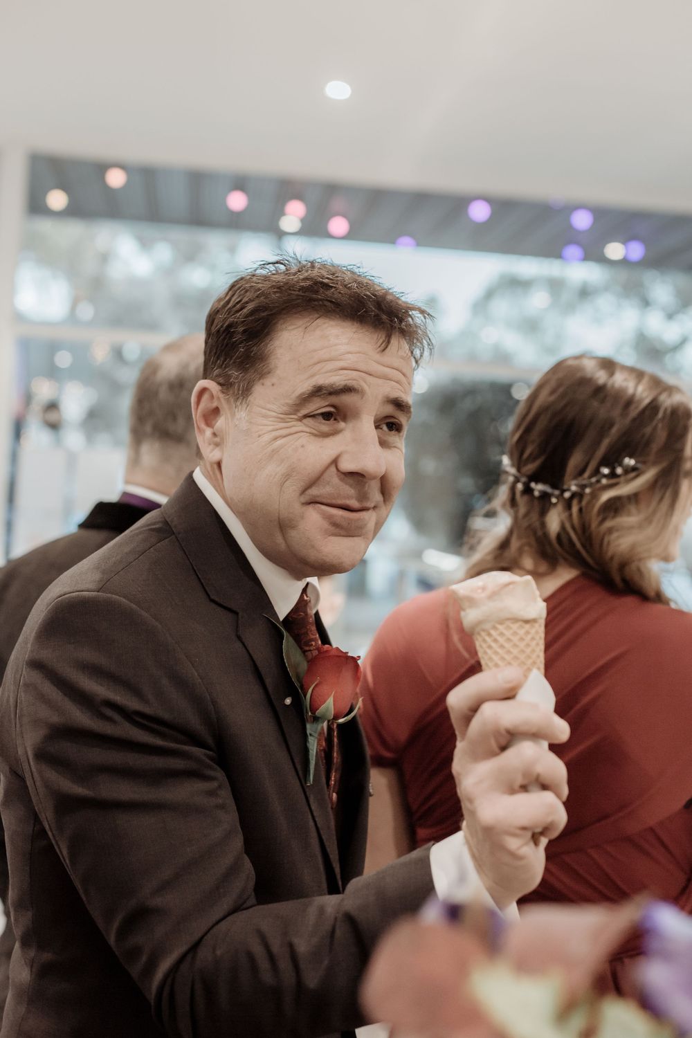 A guest in a brown suit and red tie enjoys an ice cream cone at an evening event with twinkling lights in the background.